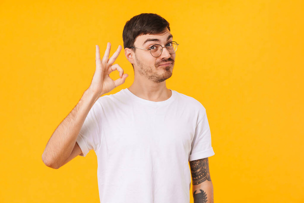 Photo of amusing young man in eyeglasses making ok sign and looking at camera isolated over yellow background - Valokuva, kuva