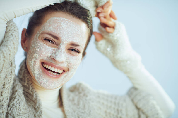 Portrait of smiling modern woman in roll neck sweater and cardigan with white facial mask as part of winter skin care isolated on winter light blue background. - Photo, Image