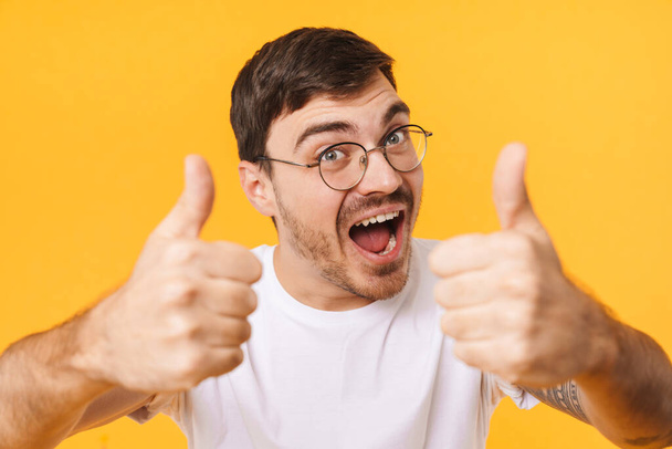 Photo closeup of joyful young man in eyeglasses showing thumbs up and smiling isolated over yellow background - Fotoğraf, Görsel