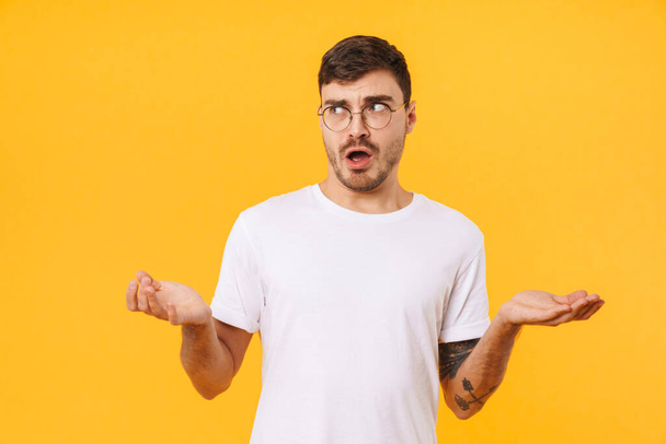 Photo of confused young man in eyeglasses posing and looking at camera isolated over yellow background - Foto, afbeelding
