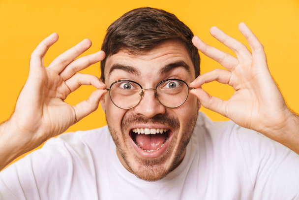 Photo closeup of excited young man in eyeglasses expressing surprise and looking at camera isolated over yellow background - Foto, Bild