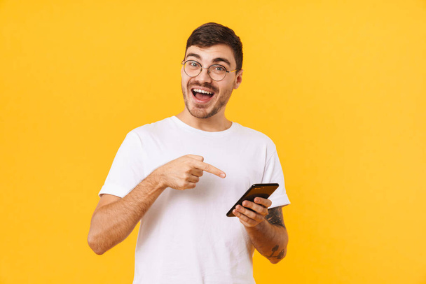 Photo of joyful young man in eyeglasses holding and pointing finger at cellphone isolated over yellow background - Photo, Image