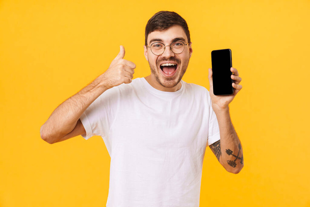 Photo of joyful young man in eyeglasses holding cellphone and showing thumb up isolated over yellow background - Fotografie, Obrázek