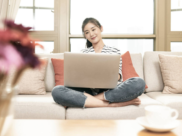 joven asiático mujer de negocios trabajo desde casa sentado en sofá usando laptop ordenador - Foto, Imagen