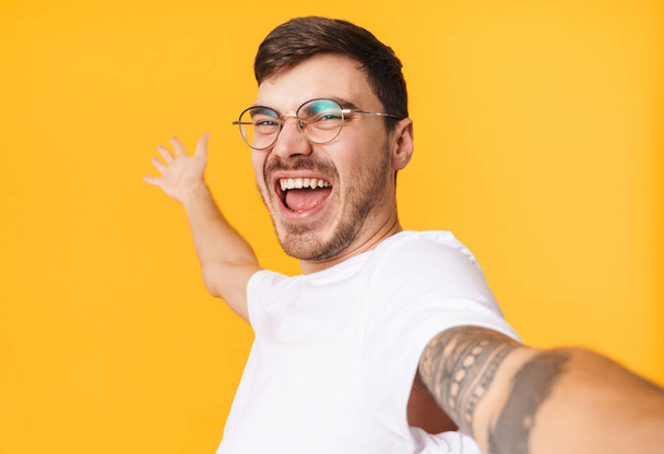 Photo of joyful young man in eyeglasses taking selfie on cellphone and smiling isolated over yellow background - Фото, изображение