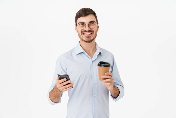 Photo of happy caucasian man in eyeglasses drinking coffee and using cellphone isolated over white background - Foto, immagini
