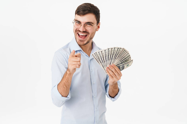 Photo of delighted man in eyeglasses holding dollars and pointing finger at camera isolated over white background - Фото, изображение
