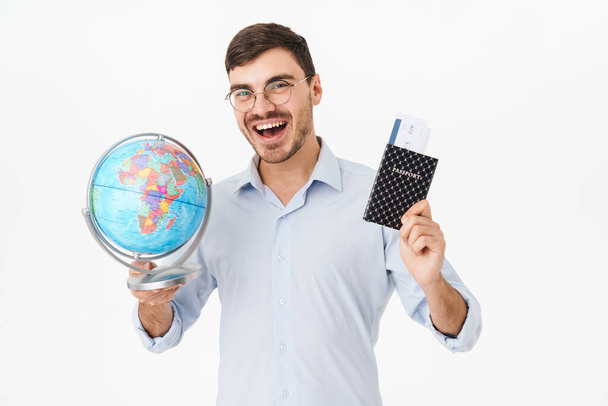 Photo of delighted man in eyeglasses holding passport with tickets and globe isolated over white background - Photo, Image