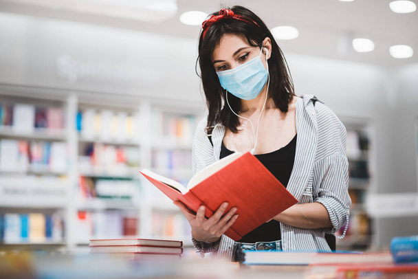 Young female student dressed in trendy outfit preparing for exam and reading a book at the library.Cute hipster girl with face medical mask looking for study references at University during pandemic - Foto, Imagem