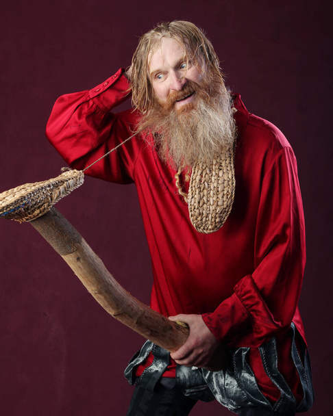 portrait of a man in a red shirt with long hair beard and mustache with a baton and bast shoes in hands studio on a burgundy background - Foto, Imagem