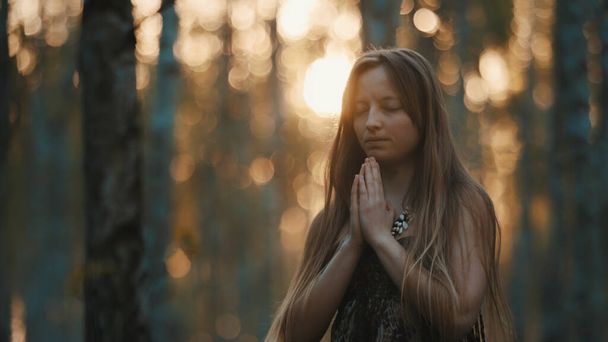 young woman praying with closed eyes in nature during the sunset surrounded by trees. - Photo, Image