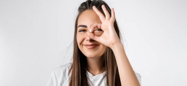 Close up portrait of carefree modern girl with long chestnut hairs, looking at the camera through fingers in OKAY gesture. Face expressions, emotions, and body language - Photo, Image