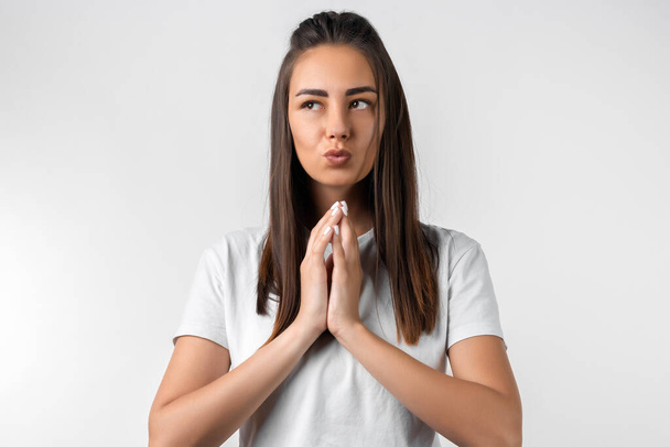 Photo of charismatic Pensive European young woman with long chestnut hair keeps hands together near chin, has cute expression, wears white t-shirt. - Фото, зображення