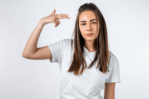 I'm fed up with this. Stylish European girl with long chestnut hair shoots in temple, tilts head, dressed in casual t shirt, demonstrates suicide gesture, isolated on white background - Photo, Image