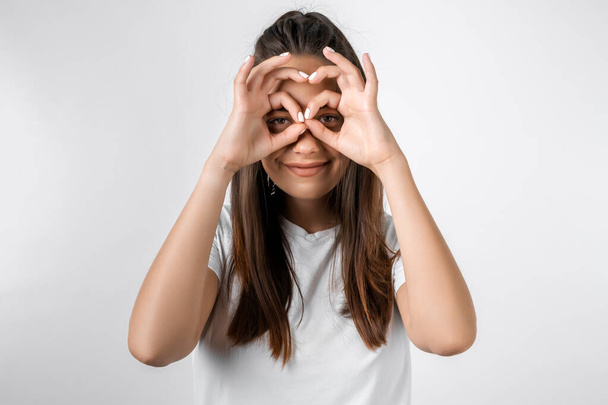 Young cheerful girl with long chestnut hairs in casual light clothes posing isolated on white wall background. People lifestyle concept. Holding hands near eyes, imitating glasses or binoculars - Φωτογραφία, εικόνα