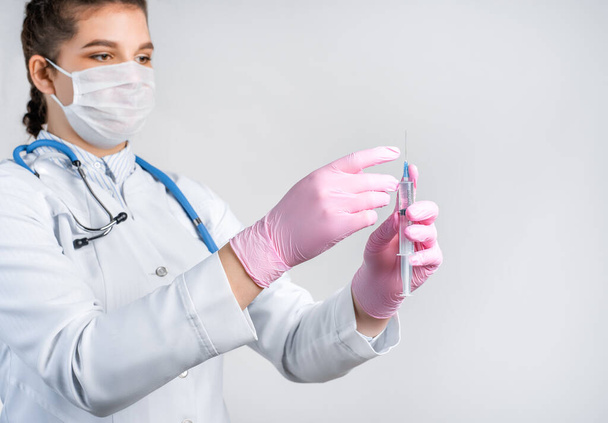 A young female doctor in a white coat, in a medical mask and pink sterile gloves hold syringe. Focus at syringe. Vaccination concept. - Photo, Image
