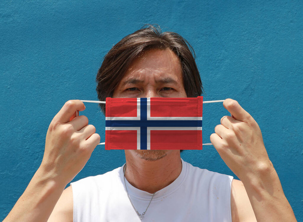 Um homem com a bandeira da Noruega em máscara higiênica na mão e levantou o rosto da frente em fundo azul. Pequena partícula ou coroa de vírus ou proteção Covid 19. Conceito de Combate à Doença. - Foto, Imagem