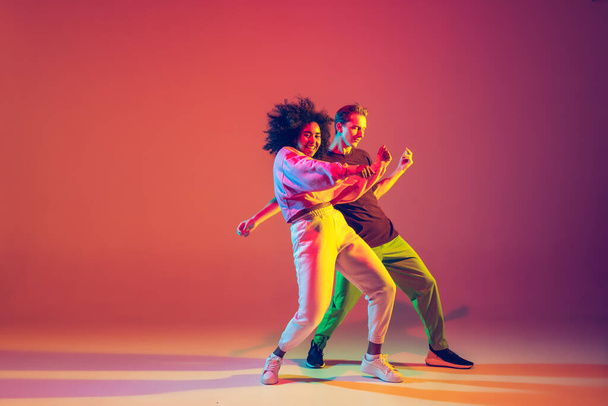 Stylish man and woman dancing hip-hop in bright clothes on gradient background at dance hall in neon light - Фото, изображение