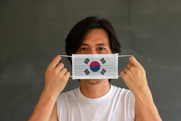 Ein Mann mit südkoreanischer Flagge auf Hygienemaske in der Hand und hochgezogenem Gesicht auf dunkelgrauem Hintergrund. Winzige Partikel oder Viren Corona oder Covid 19 Schutz. Konzept zur Bekämpfung von Krankheiten. - Foto, Bild