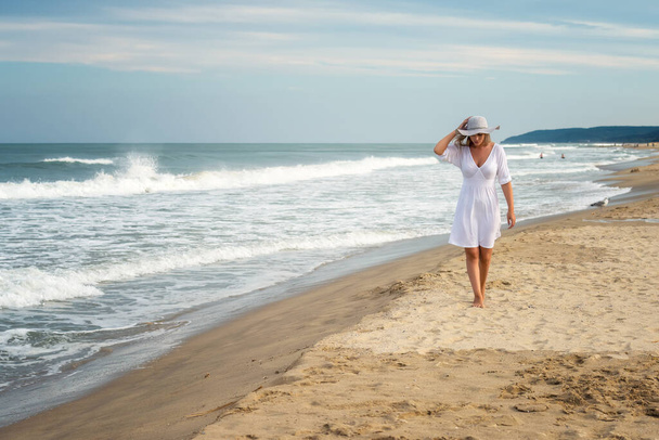 View of a beautiful young woman in white dress and summer hat walking on a sandy beach - Photo, image