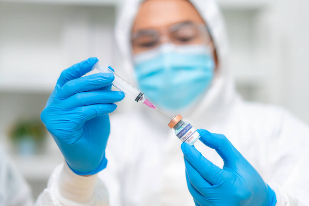 Bliska dłoń lekarza lub naukowca w mundurze zestawu PPE noszącego maskę ochronną w butelce i strzykawce zawierającej szczepionkę zawierającą płyn, koronawirus lub białą koncepcję COVID-19 - Zdjęcie, obraz