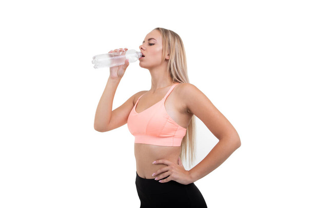 Egy sportos szőke lány rózsaszín felsőben vizet iszik egy üvegből. - Fotó, kép