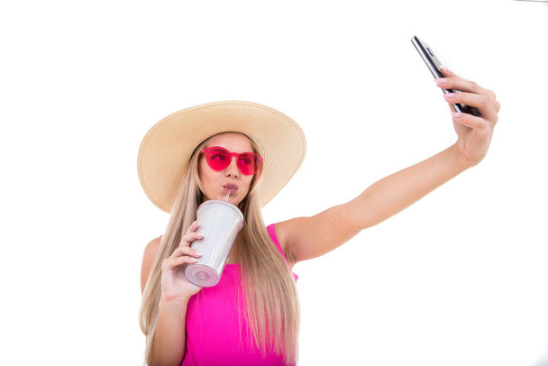Dziewczyna w różowym stroju kąpielowym i kapeluszu trzymająca szklankę i robiąca selfie. Izolacja - Zdjęcie, obraz