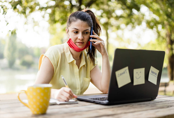 Young woman wearing lowered protective face mask using laptop computer outdoors - Female freelancer or manager working on laptop at park - Remote working and new normal concept during coronavirus time - Photo, Image