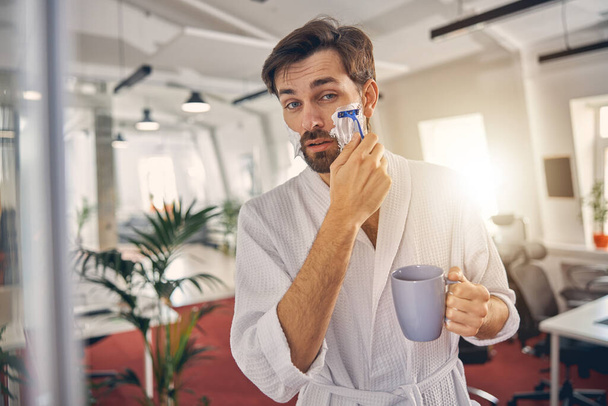 Tired young man in bathrobe shaving at work - Photo, Image