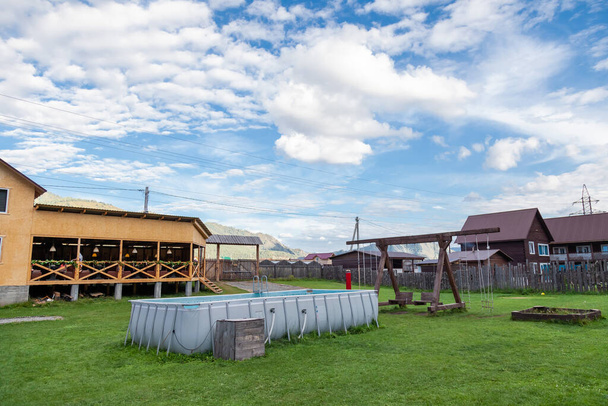 Um grande balanço feito de madeira e uma piscina de borracha inflável em um gramado de grama verde no pátio de uma casa de campo com uma cerca de madeira contra o pano de fundo da montanha e céu azul. - Foto, Imagem