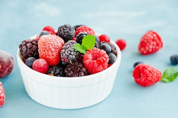 Mix of summer frozen berries in a white bowl on a blue concrete background. Copy space - Zdjęcie, obraz