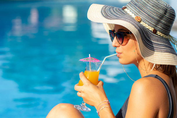 Close up face shot of cute woman wearing sun hat enjoying fruit cocktail at resort pool. - Zdjęcie, obraz