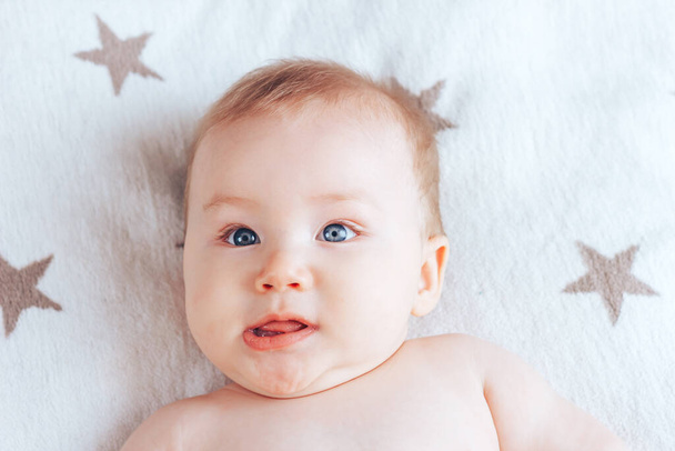 カメラには赤ん坊が顔を出し、青い目をした新生児と、茶色の星のついたライトブランケットの上にブロンドの髪が横たわっています。. - 写真・画像