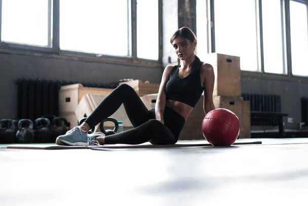 Young woman sitting on floor after her workout and looking down. Female athlete taking rest after fitness training - Photo, Image