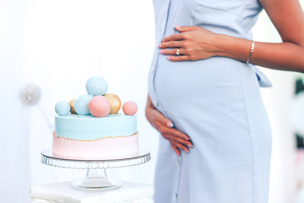 Giovane mamma incinta in un abito blu sta toccando la pancia in piedi accanto a una bella torta. - Foto, immagini