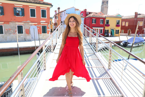 Νεαρή όμορφη γυναίκα με κόκκινο φόρεμα περπατώντας στη γέφυρα στο χωριό Burano, Βενετία, Ιταλία - Φωτογραφία, εικόνα