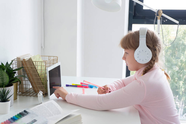 teen girl si siede a un tavolo con un tablet e in cuffia e scrive in un notebook. concetto di apprendimento a distanza. - Foto, immagini