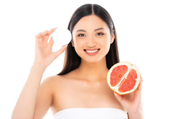 brunette asian woman looking at camera while holding half of juicy grapefruit and cosmetic cream isolated on white - Photo, Image