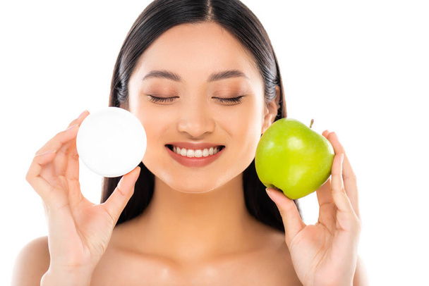 mujer asiática desnuda con los ojos cerrados sosteniendo toda la manzana verde y crema cosmética cerca de la cara aislada en blanco - Foto, imagen