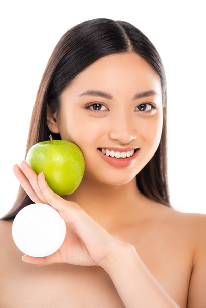 mujer asiática desnuda sosteniendo manzana verde madura y crema cosmética cerca de la cara aislada en blanco - Foto, imagen