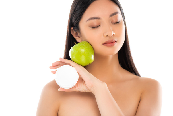 nahá asijská žena se zavřenýma očima drží zralé jablko a kosmetický krém v blízkosti obličeje izolované na bílém - Fotografie, Obrázek