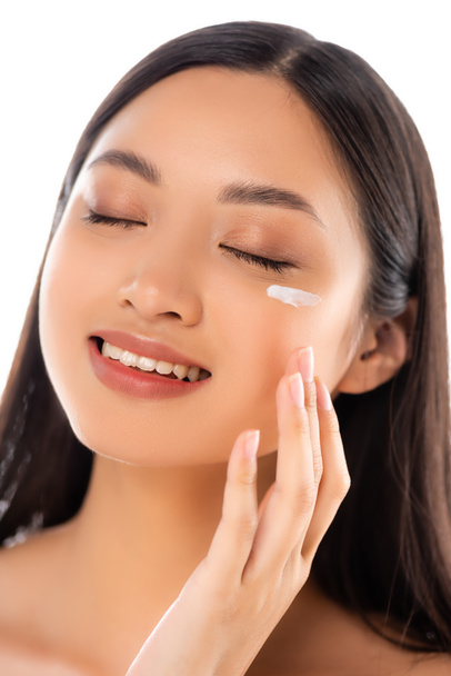 brunette asian woman with closed eyes applying cosmetic cream on face isolated on white - Foto, Bild