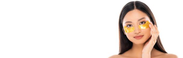 immagine orizzontale di donna asiatica toccare patch occhio dorato sul viso mentre guardando la fotocamera isolata su bianco - Foto, immagini