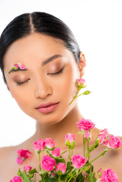 Yüzünde tomurcuklar ve taç yaprakları olan genç Asyalı bir kadın beyaz güllerin yanında gözlerini kapatıyor. - Fotoğraf, Görsel