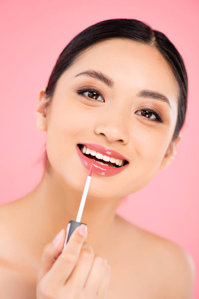 brunette asian woman looking at camera while applying lip gloss isolated on pink - Foto, immagini