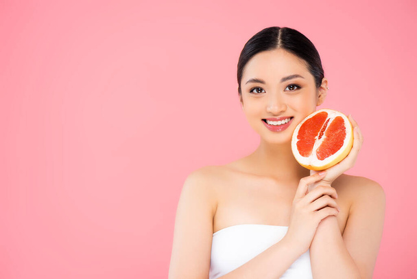young asian woman holding half of grapefruit near face and looking at camera isolated on pink - Photo, Image