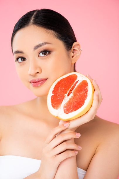 young asian woman holding half of ripe grapefruit while looking at camera isolated on pink - Foto, Bild