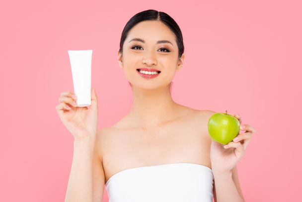 aziatische vrouw met cosmetische crème en groene appel terwijl kijken naar camera geïsoleerd op roze - Foto, afbeelding