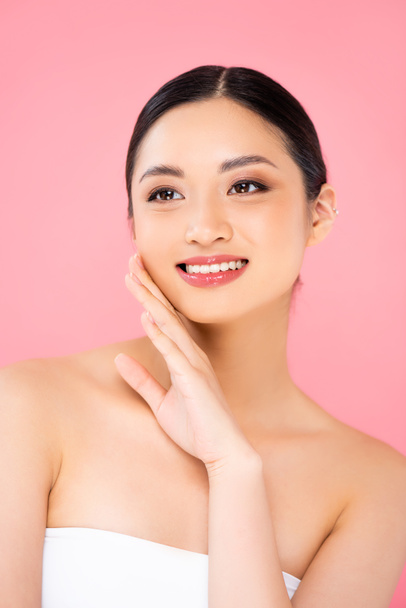 brunette asian woman looking away while touching face isolated on pink - Фото, изображение