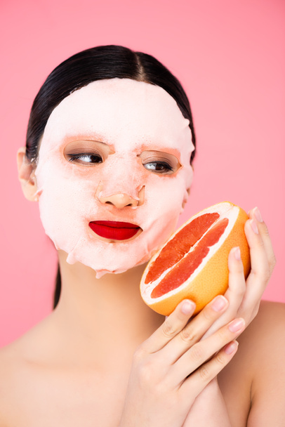 morena asiática mujer en la cara máscara celebración de la mitad de jugoso pomelo aislado en rosa - Foto, Imagen